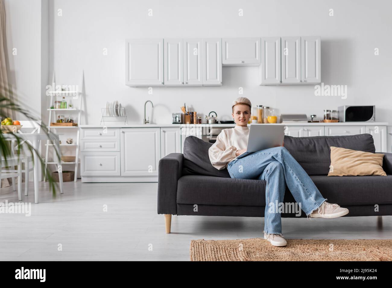 full length of woman in jeans sitting on couch with laptop in modern ...