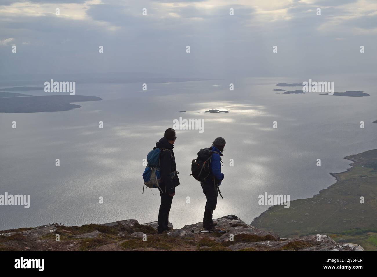 Hikers/hill walkers in silhouette on Ben Mor Coigach, a ridge near Ullapool, north west Scotland, Assynt, admire views of Summer Isles on a spring day Stock Photo