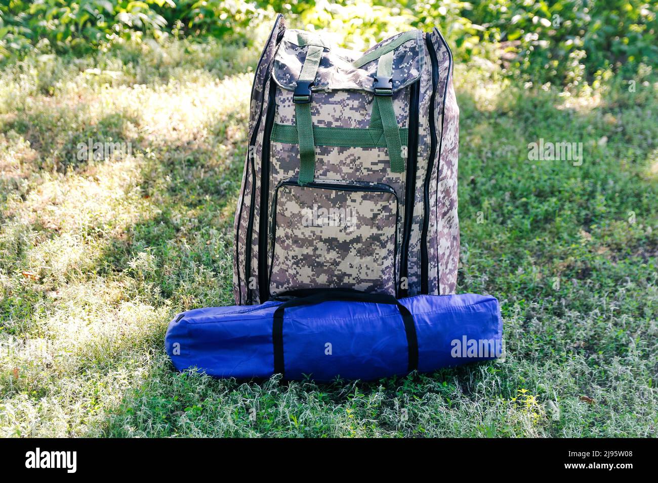 Defocus military backpack and blue tent or sleeping bag. Army bag on green grass background near tree. Military camouflage army rucksack. Vertical. To Stock Photo