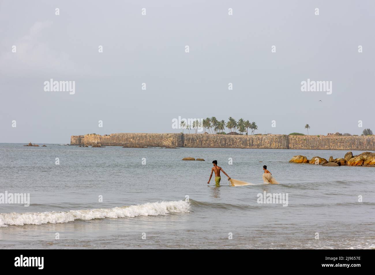 Two fishermen venturing into the sea to lay fishing nets while the massive stone wall of Sindhudurg Fort is seen in the background Stock Photo