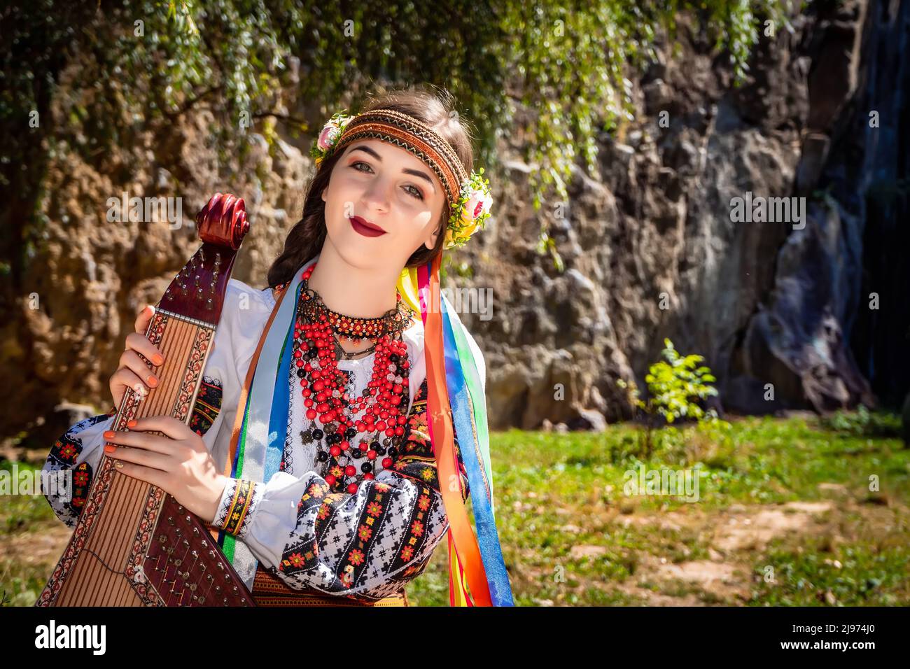 Ukrainian musician in authentic national dress sits by a rock. Ukrainian woman with a bandura musical instrument before performing folk music Stock Photo