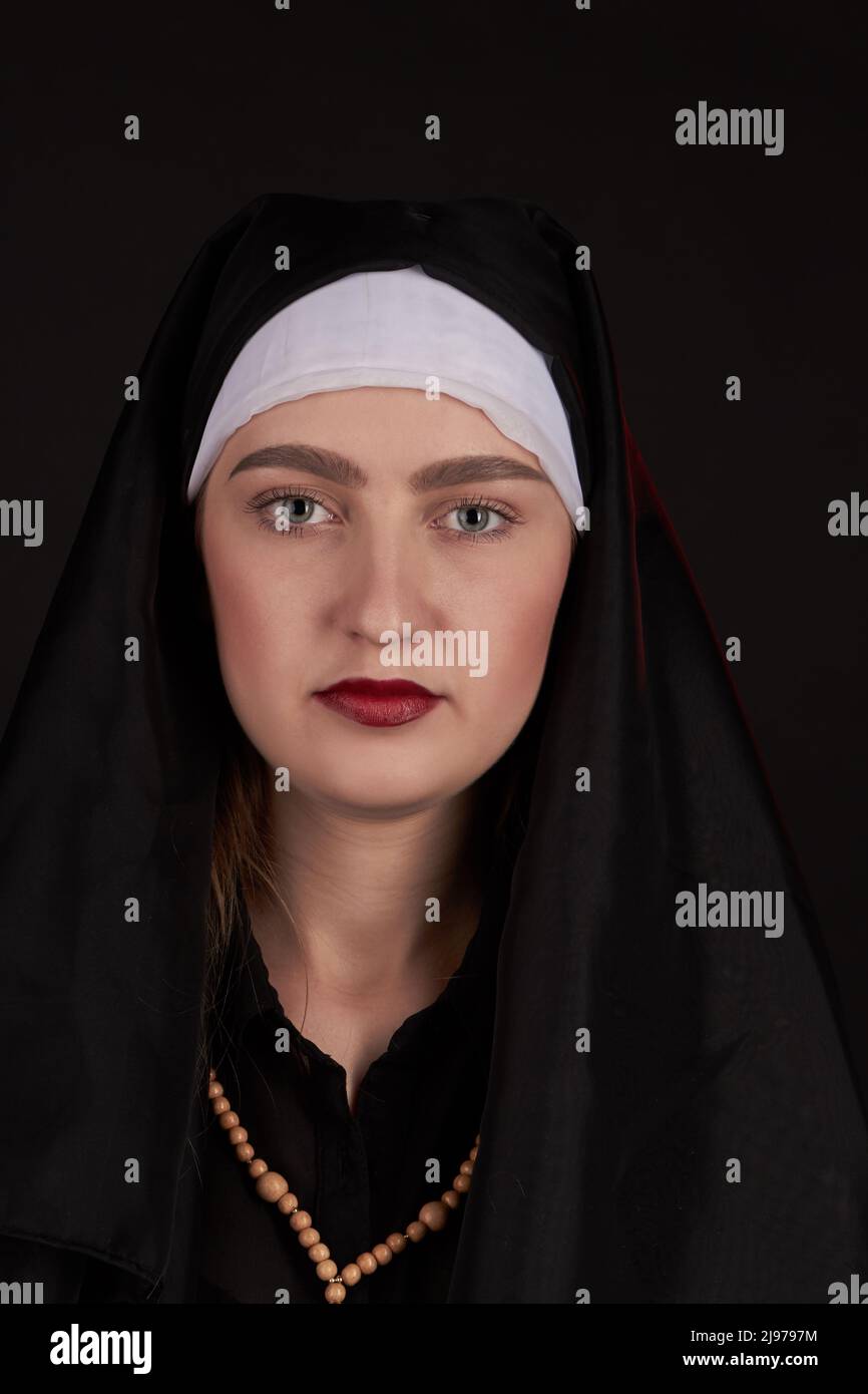 Front portrait of the young beautiful nun isoalted on black background. Vertical shot caucasian woman face wearing dark clothes. Stock Photo