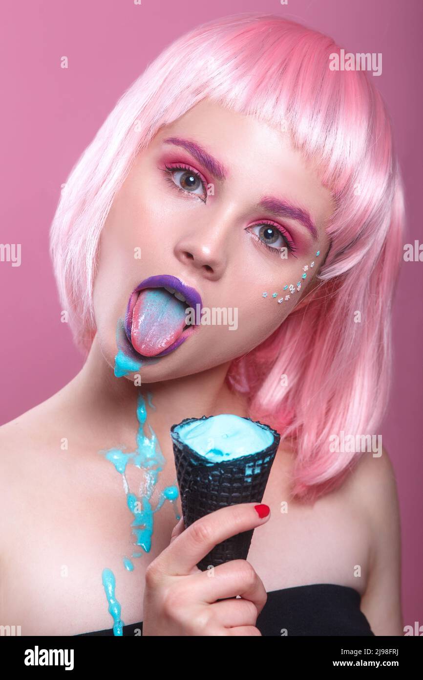 sexy young woman with pink bob cut and purple lips eating blue ice cream in black waffle glass on pink background Stock Photo