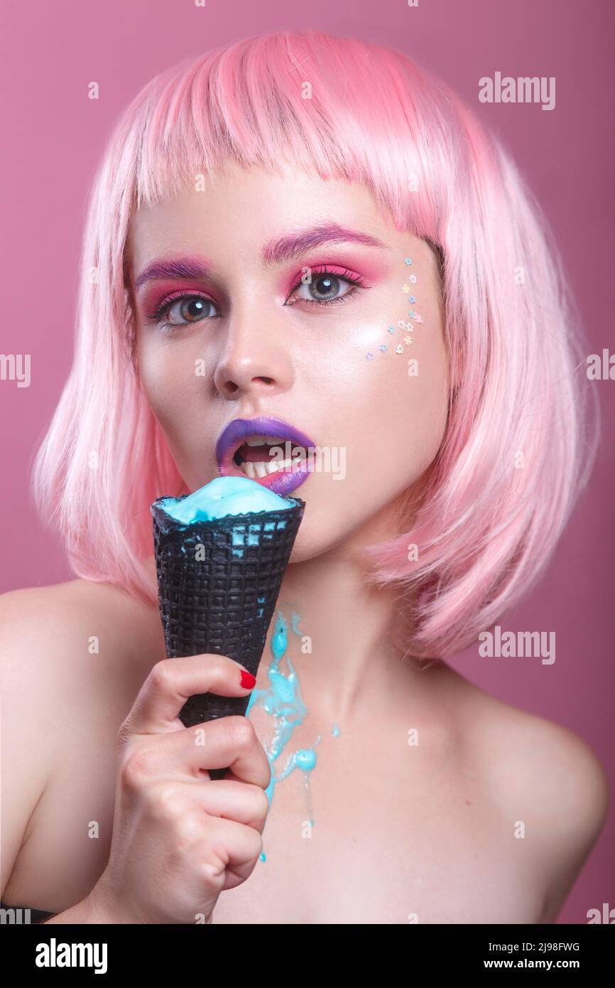 sexy young woman with pink bob cut and purple lips eating blue ice cream in black waffle glass on pink background Stock Photo