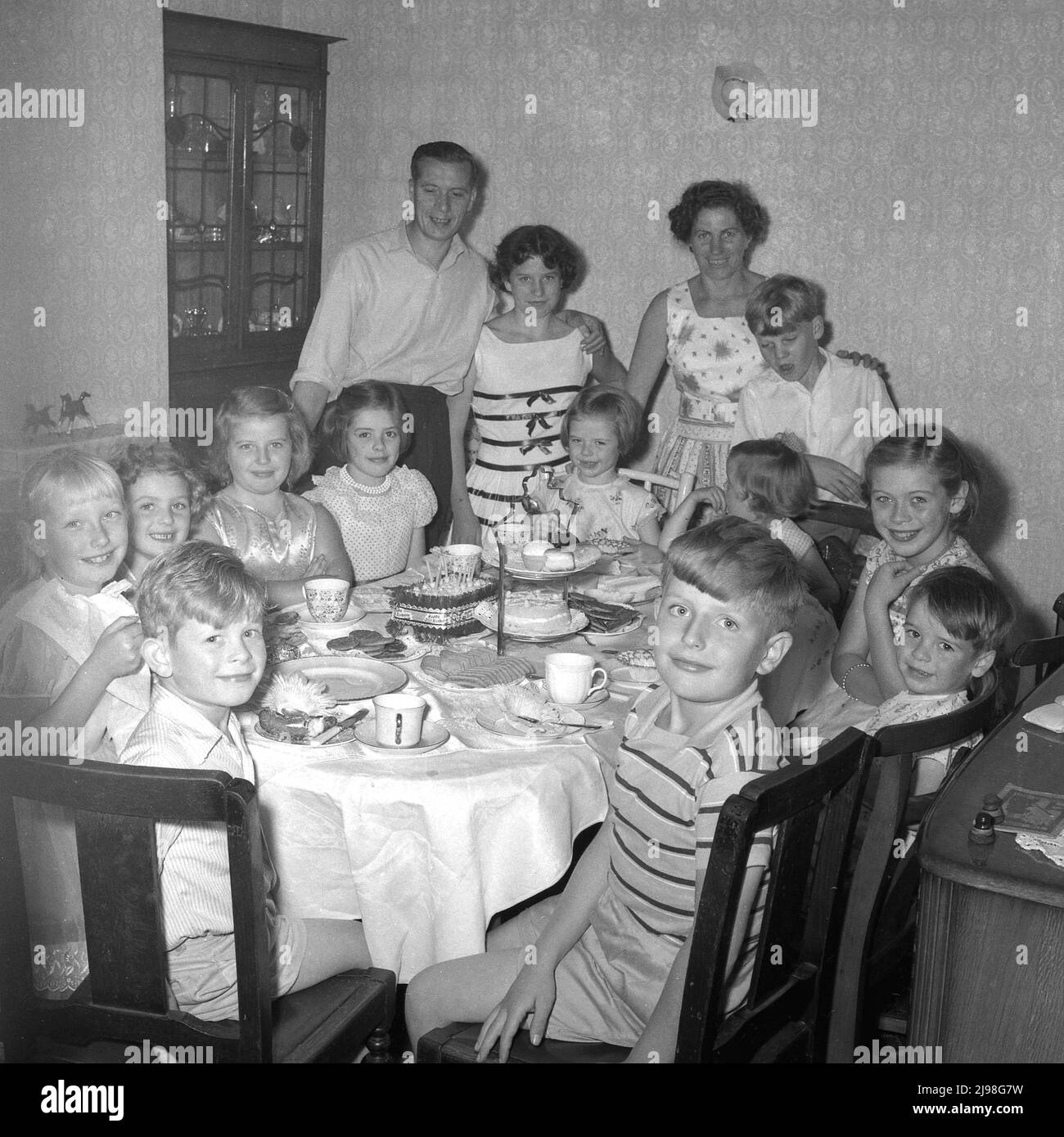 1961, historical, birthday, lovely smiles from a group of children sitting around a table enjoying a celebatory birthday tea party, with mum and dad standing by for a group photo, England, UK. Stock Photo