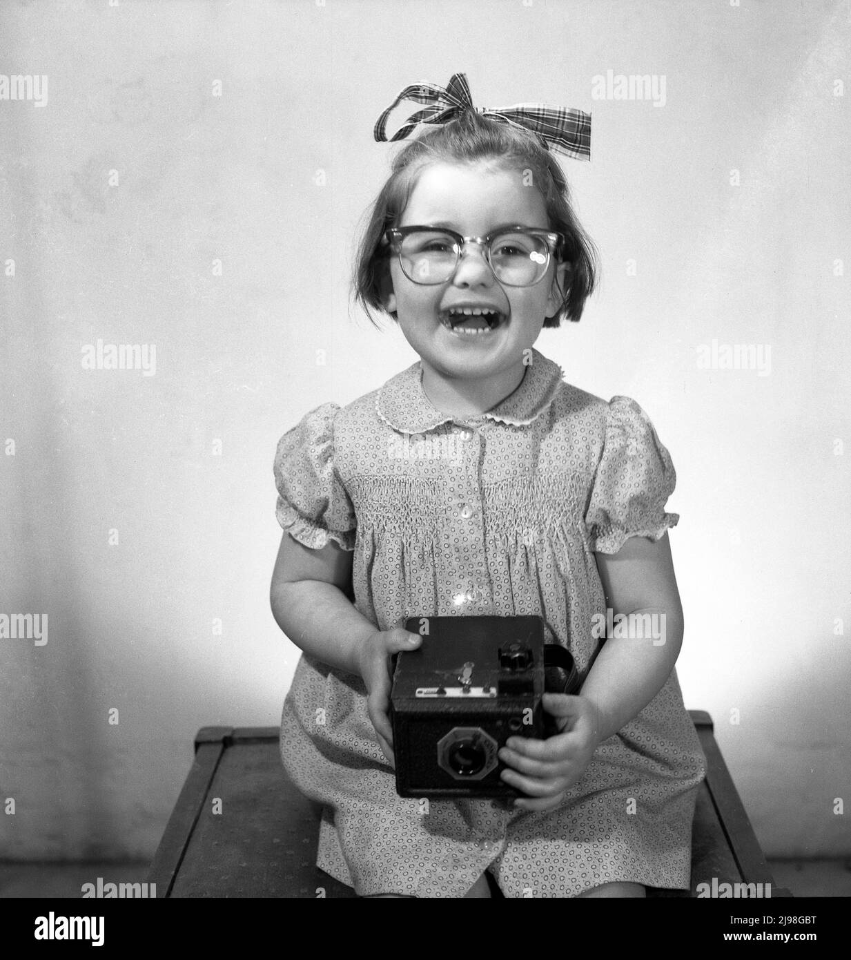 1963, historical, studio photo, a happy young girl with a big smile and with a ribbon in her hair sitting on a stool for her picture, wearing her mother's glasses and holding a camera of the era, a Conway box film camera, England, UK. Stock Photo