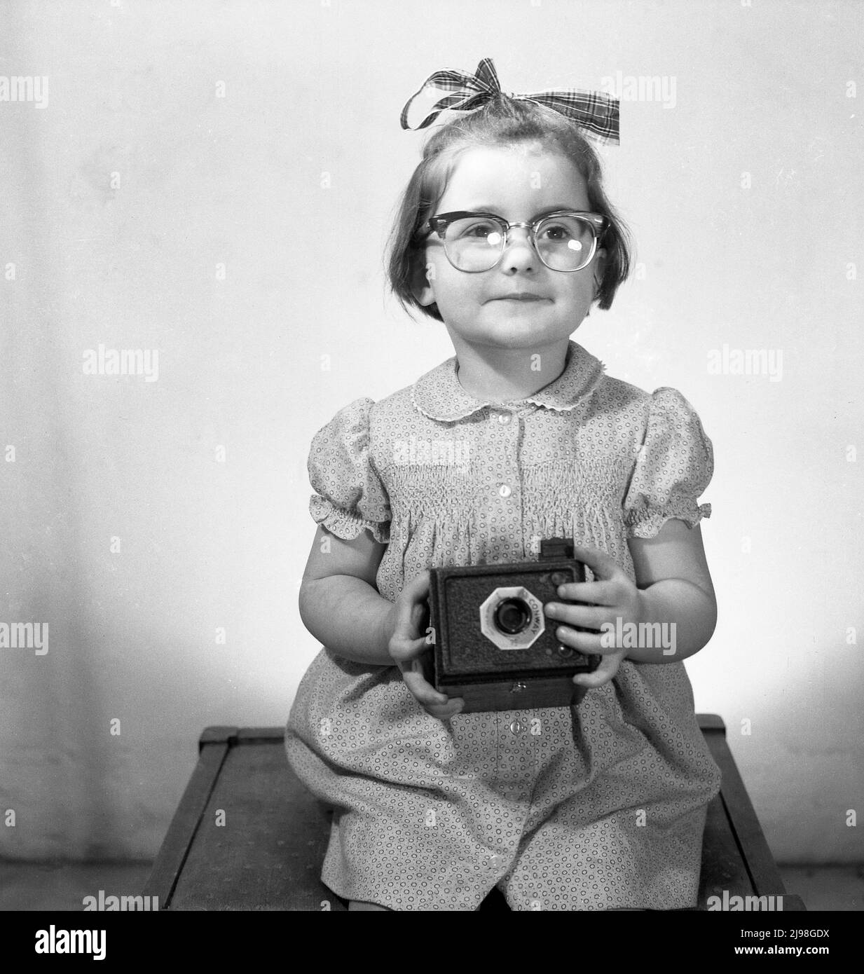 1963, historical, studio photo, a youg girl with a ribbon in her hair sitting on a stool for her picture, wearing her mother's glassses and holding a Conway box film camera, England, UK. Stock Photo