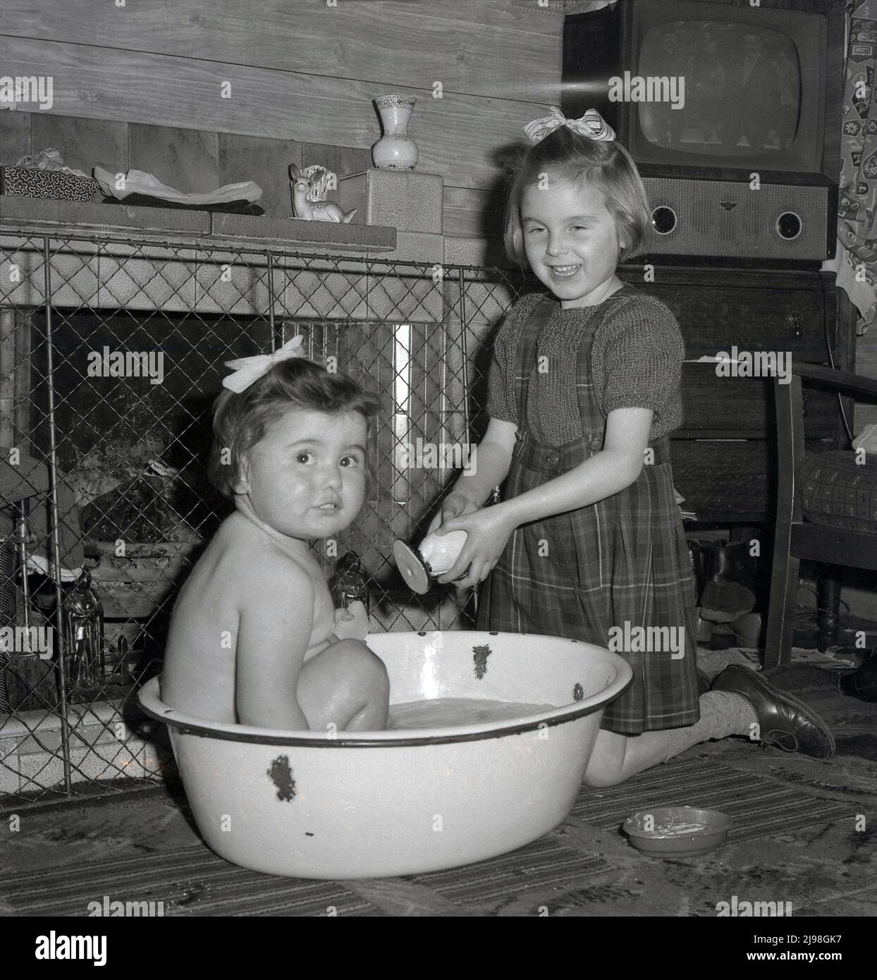 1961, historicsl, litting sitting in an enamal basin or bowl in front of the fire having a bath, with her big sister  beside her a toy penguin, Stockport, England, UK. A television set of era is in the corner of the room. Stock Photo
