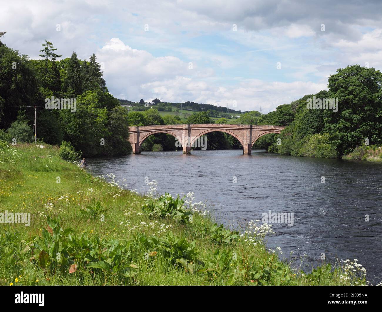 A fisherman on the grass banks of the River Beauly. The stone built Lovat Bridge (1811-1814) which carries the A862 road to Inverness. Stock Photo