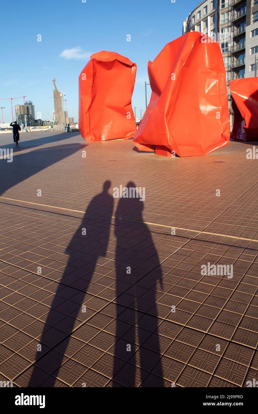 Long shadows of two people in front of the Red 'Rock Strangers' Sculpture at the harbour's mouth in Ostend Belgium Stock Photo