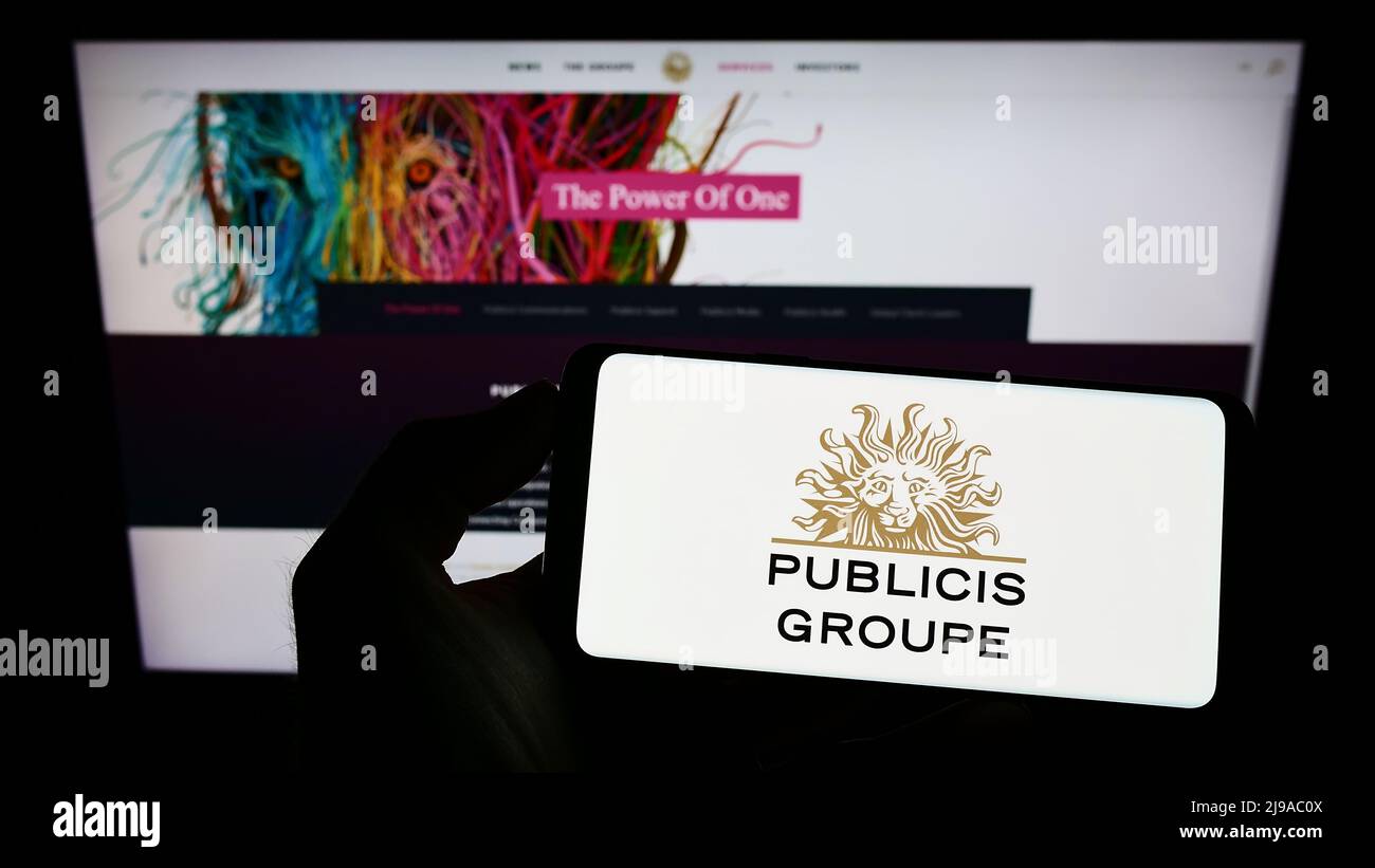 Person holding cellphone with logo of French advertising company Publicis Groupe S.A. on screen in front of webpage. Focus on phone display. Stock Photo