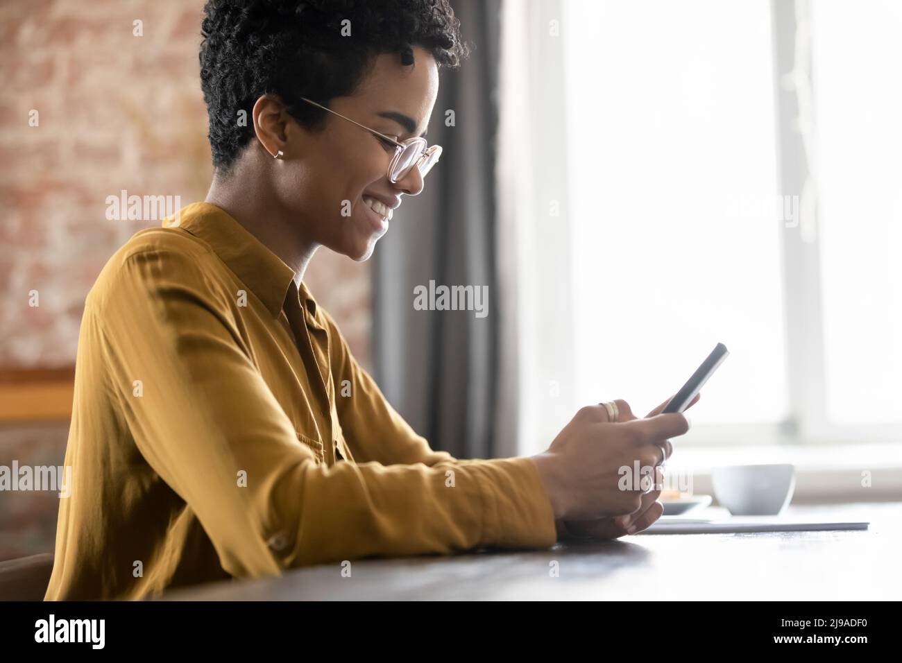 Smiling African woman in eyeglasses sit at table using smartphone Stock Photo