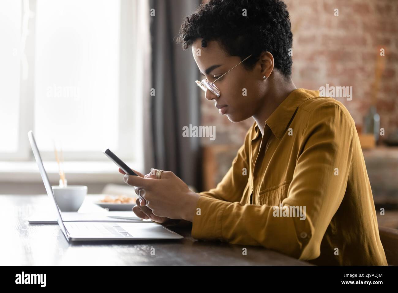 Serious African woman sit at desk with laptop using smartphone Stock Photo