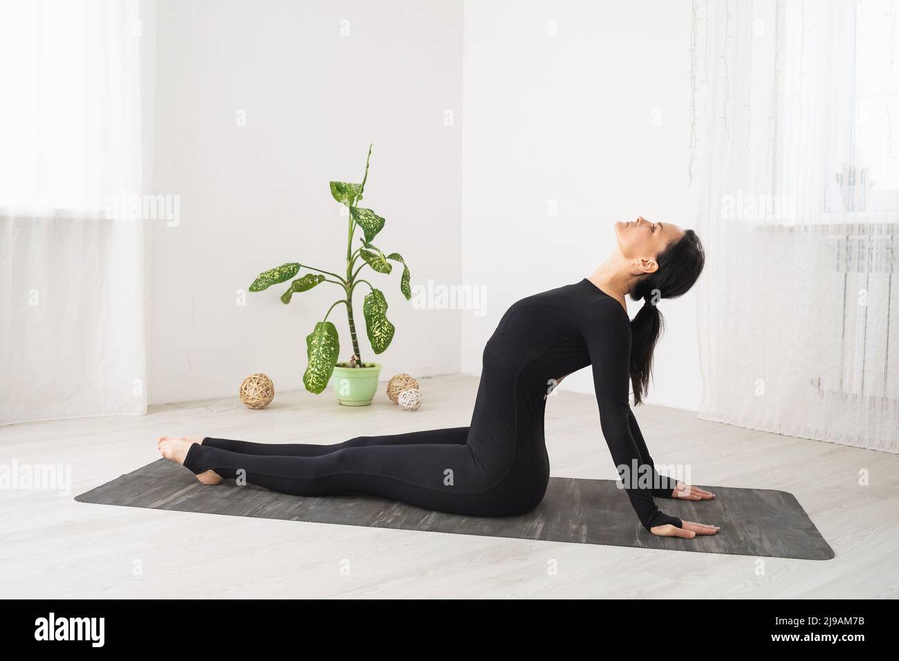 A woman in black sportswear does yoga, performs a backward deflection, trains in a room sitting on a mat Stock Photo