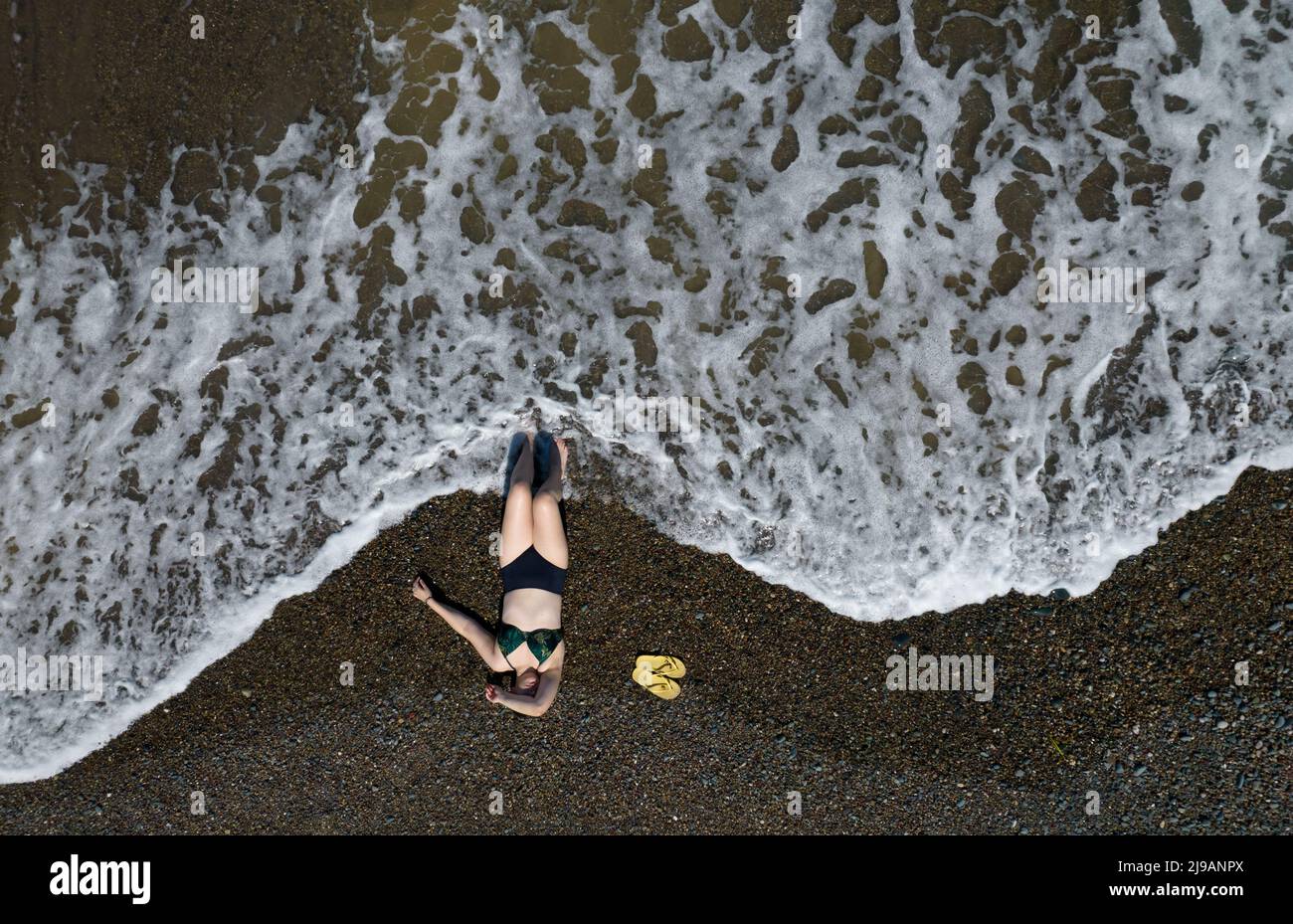 Woman with swimsuit relaxing on a sandy beach with sea waves crashing on the shore. Overhead shot. Aerial drone photograph Stock Photo