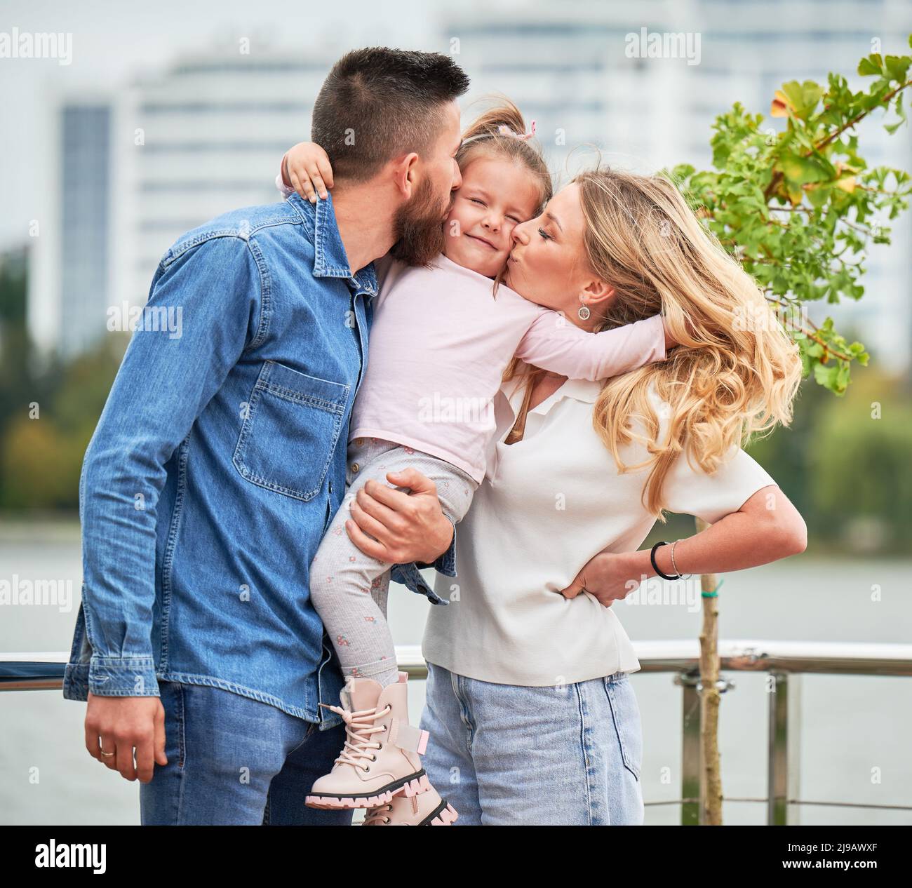 Man and woman standing on the street and kissing little girl in cheeks. Father holding adorable child and placing kiss on kid cheek while spending time with family outdoors in new urban district. Stock Photo