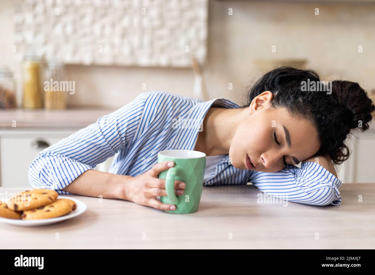 Tired latin woman with coffee cup, waking up early and sleeping on table in light kitchen interior, free space Stock Photo
