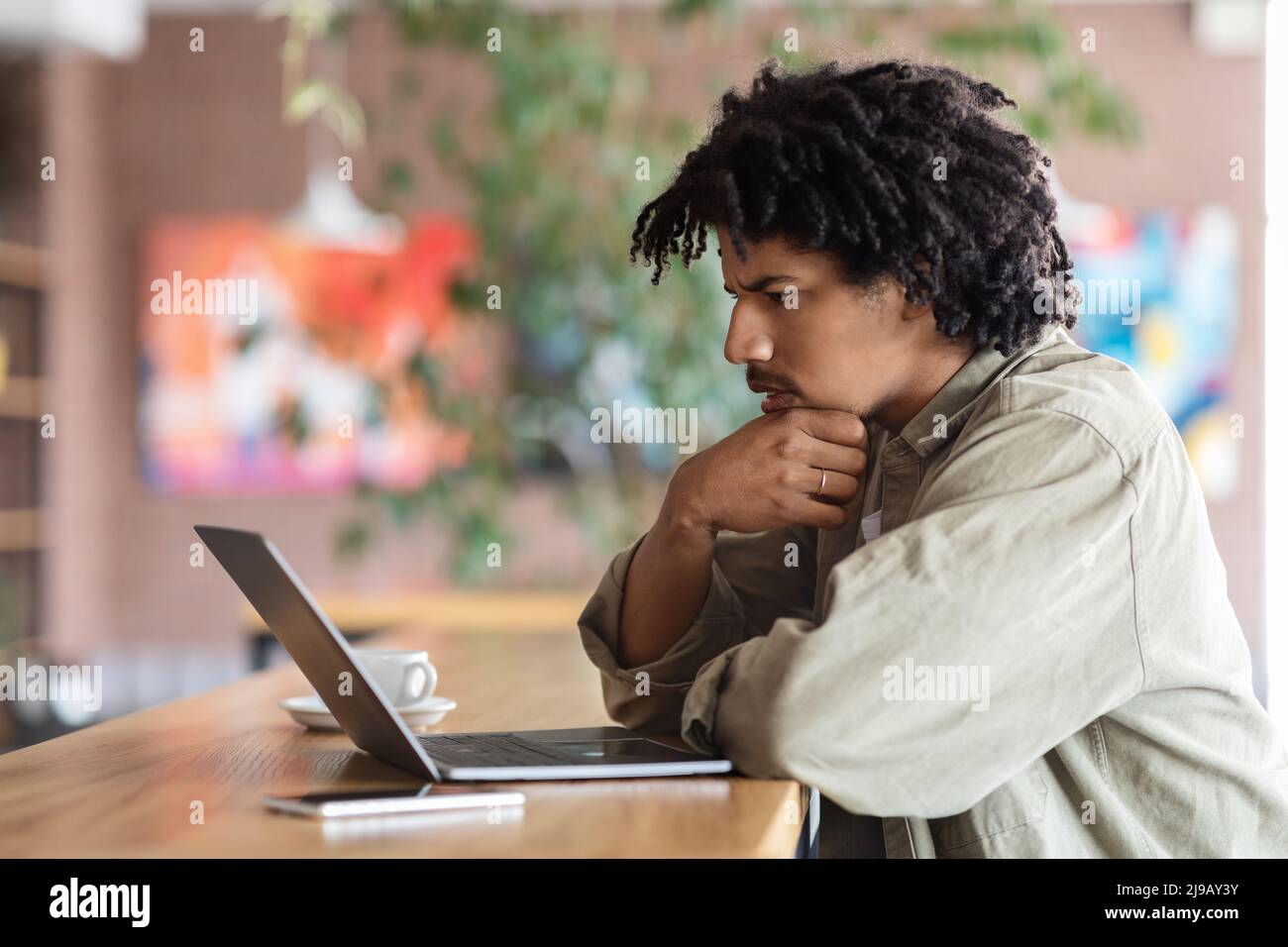 Pensive curly young black man looks at laptop screen at table in cafe interior Stock Photo