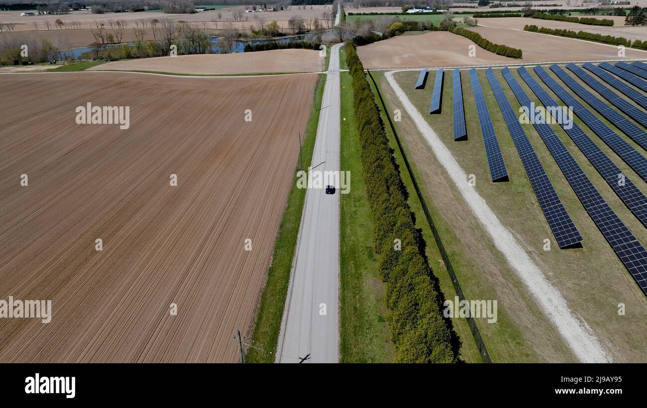 An aerial view following a blue car driving on a country road between a solar farm and field, seen on a sunny day. Stock Photo
