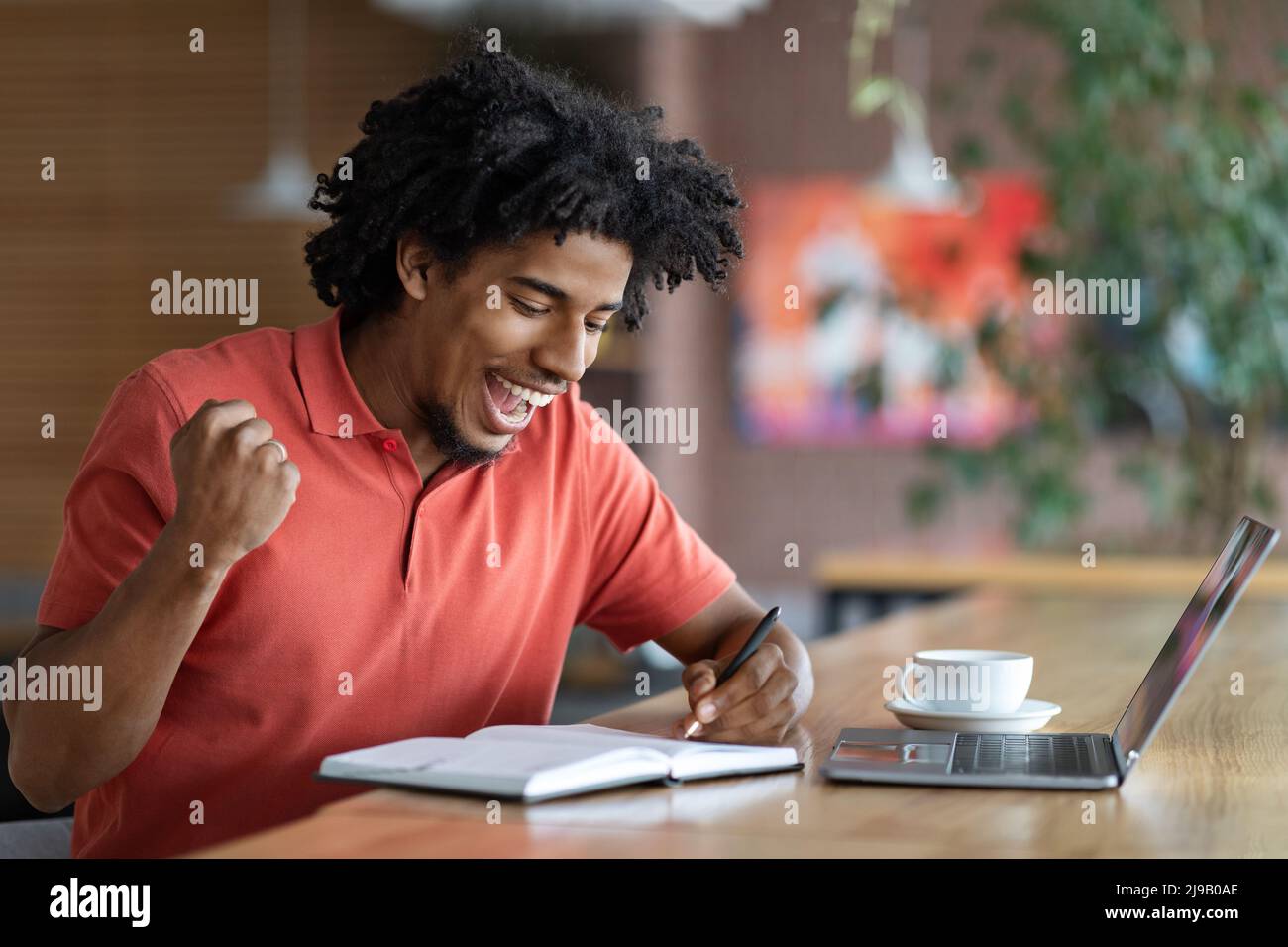 Glad happy excited curly handsome young black man make notes in notebook, victory gesture in cafe interior Stock Photo
