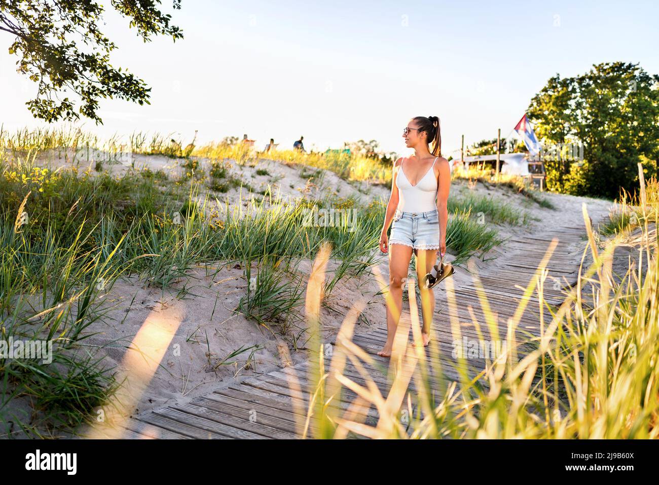 Summer sunset walk on beach boardwalk, green grass field and sand dunes. Outdoor nature portrait of happy pretty girl. Stock Photo