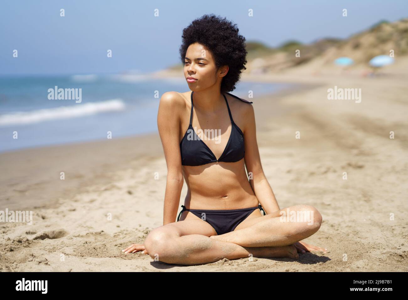 Relaxed black woman in bikini sitting on the sand of a tropical beach looking out to sea. Stock Photo