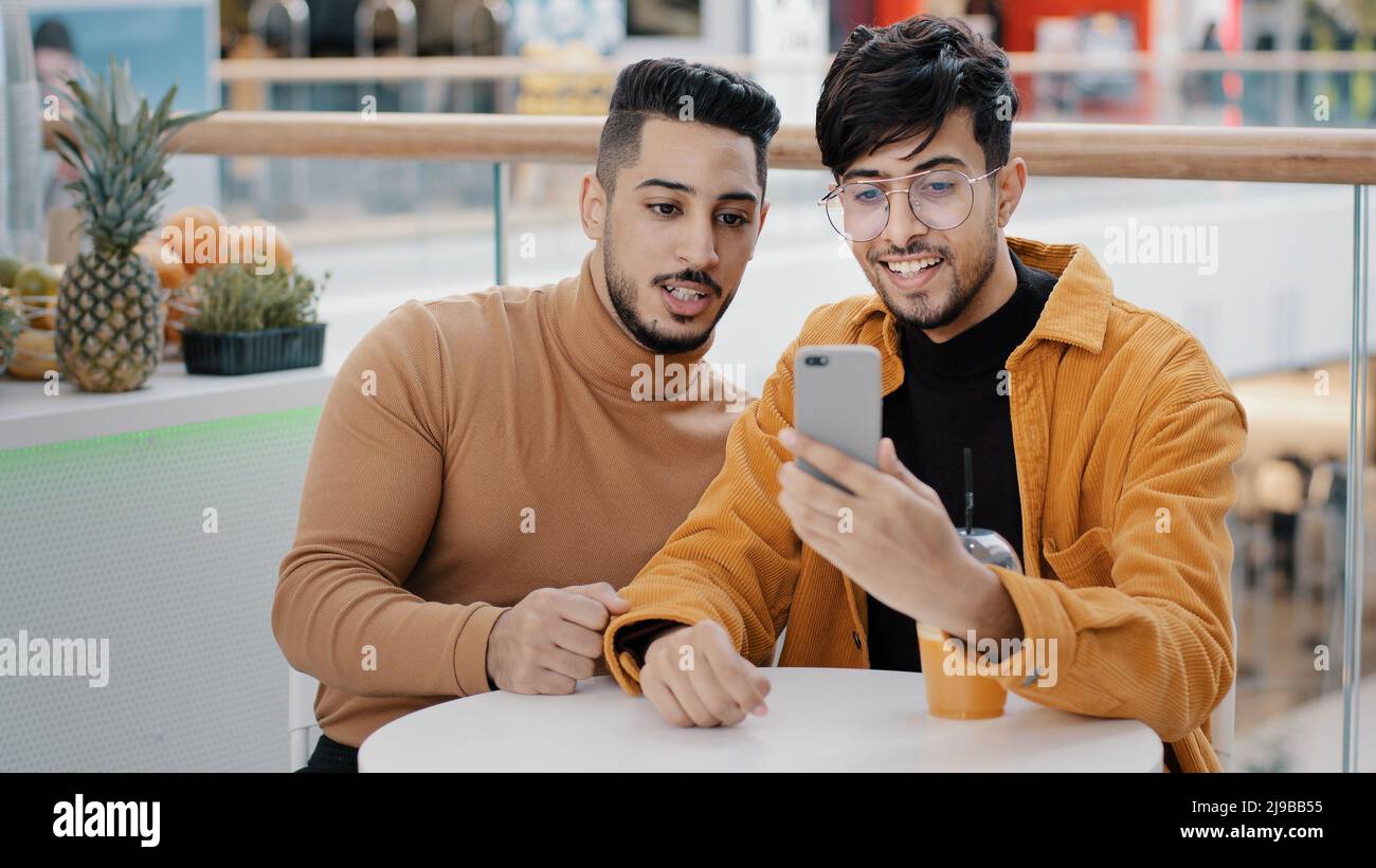 Two happy arab friend bloggers sitting in cafe recording video blog on phone communicating via webcam looking at smartphone screen take selfie making Stock Photo