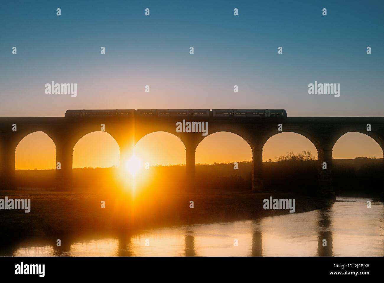 A Northern Rail train travelling over Arthington Viaduct railway bridge at sunrise with the River Wharfe beneath it. Sun rises between the arches. Stock Photo