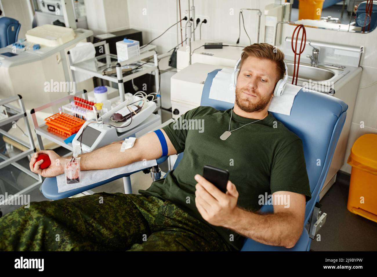 High angle portrait of military man giving blood while laying in chair at blood donation center and watching videos at smartphone, copy space Stock Photo