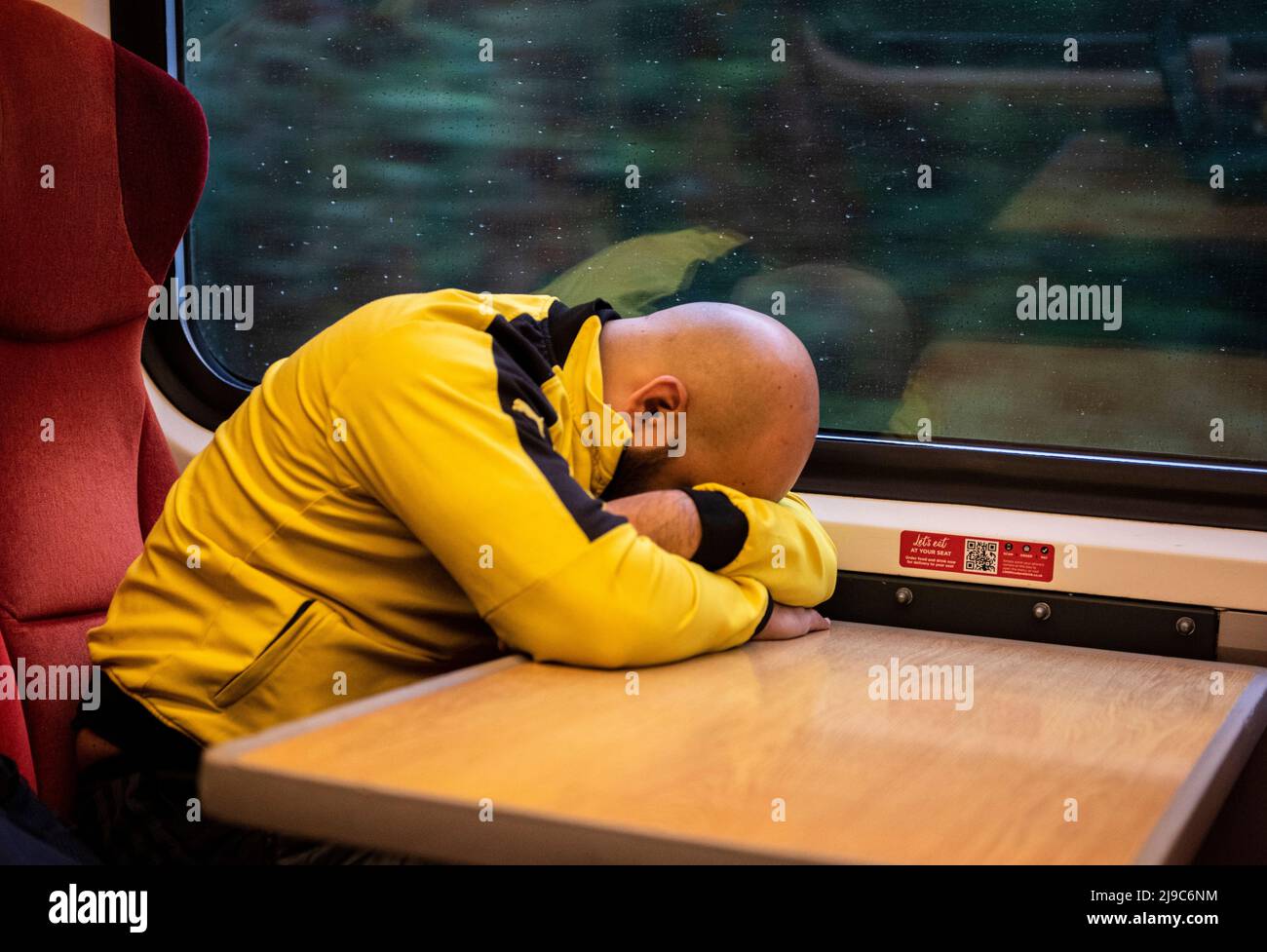 A completely bald man wearing a bright yellow top is seen asleep on a table on a train near Leeds in England. Stock Photo