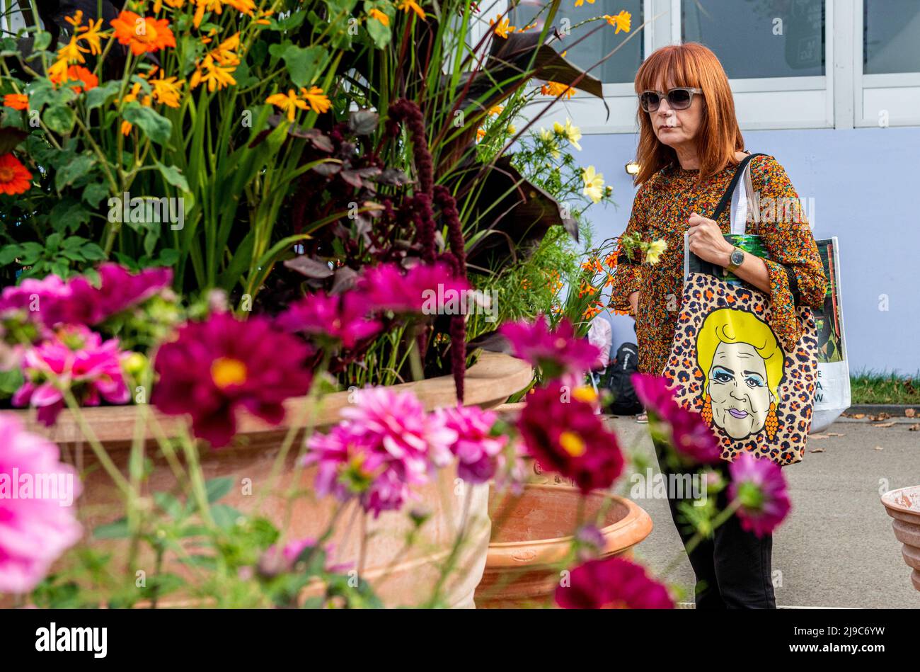 A woman wearing a floral pattern top and carrying a colourful printed bag passes on front of a big flower arrangement on the last day of the RHS Chelsea Flower Show. Stock Photo