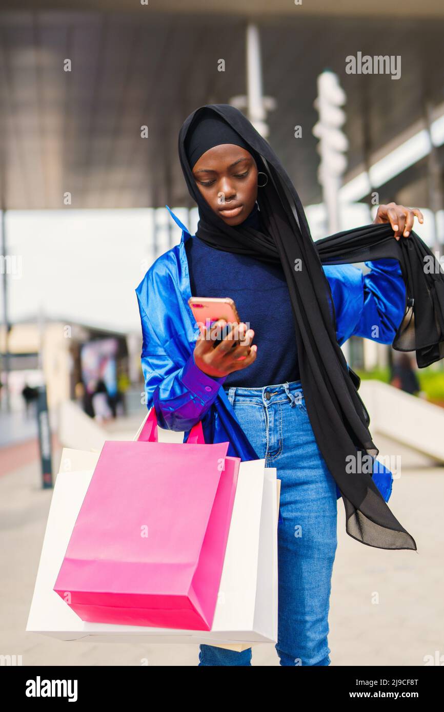 Black shopaholic adjusting hijab and using smartphone Stock Photo