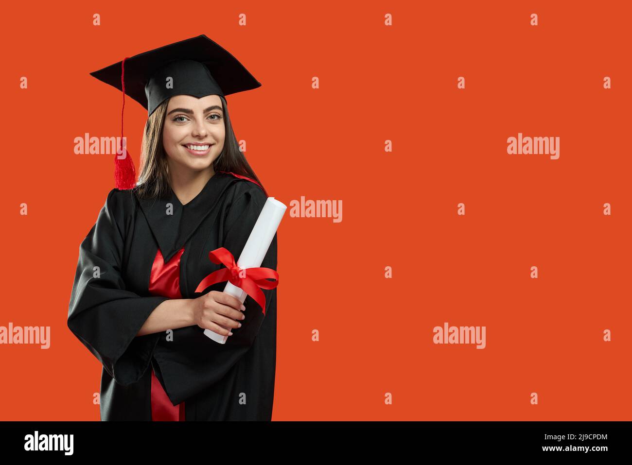 Front view of brunette girl standing with crossed hands, holding diploma. Pretty young woman graduating from college, university, high school. Isolated on red studio background.  Stock Photo