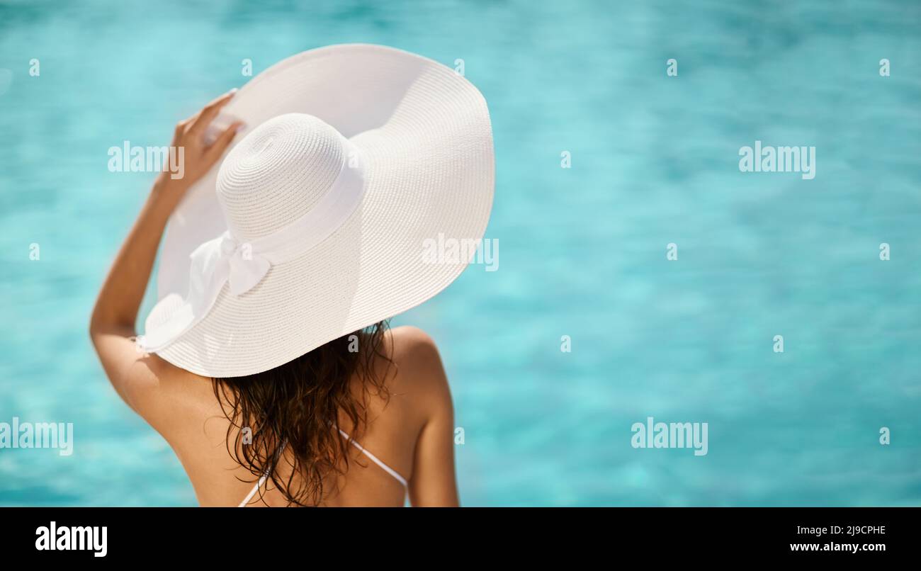 Elegant long haired woman holding panama hat against seaside at hot sunny day. Back view of brunette woman wearing big white hat looking at horizon outdoors. Concept of vacation. Stock Photo