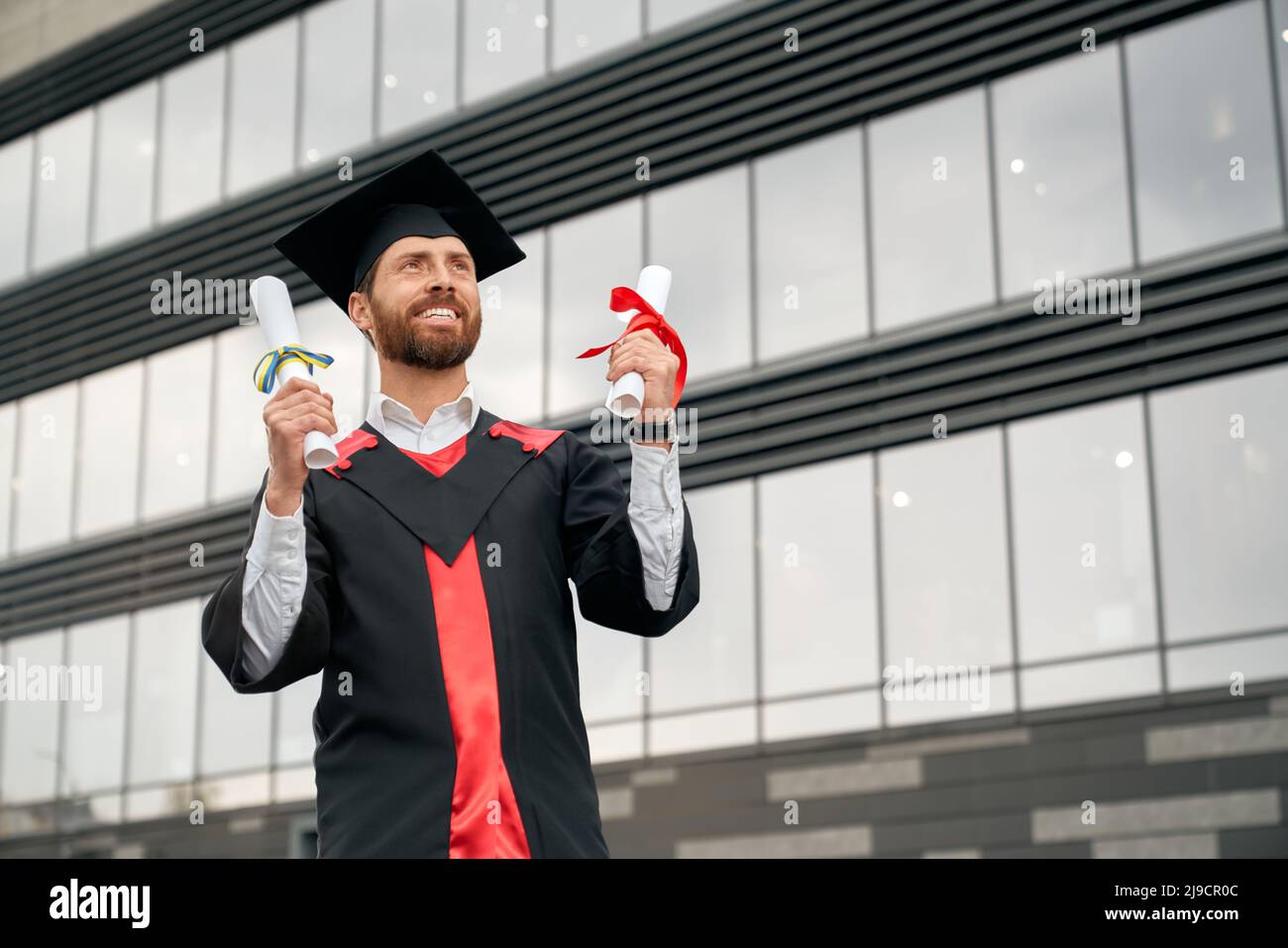 Front view of master graduating from college, university, high school. Male student in graduate gown and mortarboard standing, holding two diplomas, smiling. Concept of success. Stock Photo