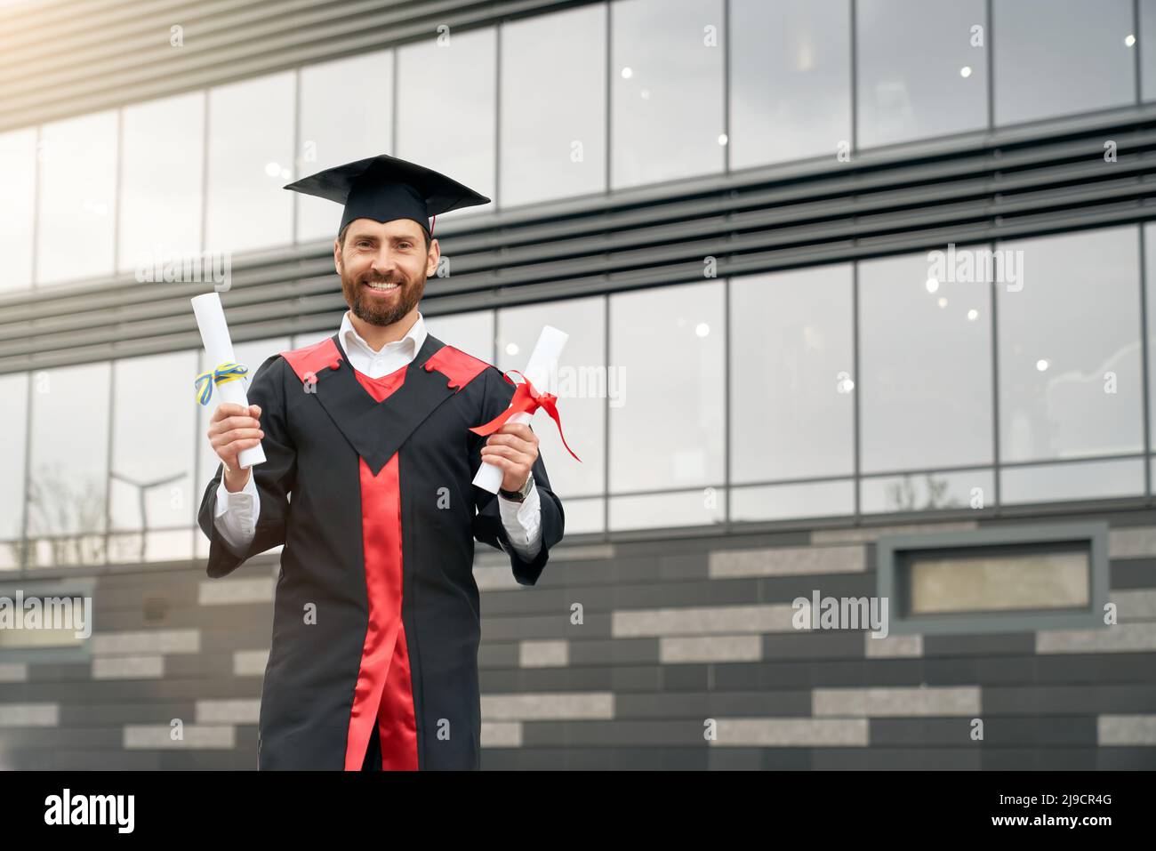 Front view of student with master degree standing, holding two diplomas, smiling. Handsome male in mortarboard and graduate gown graduating from high school. Concept of youth. Stock Photo