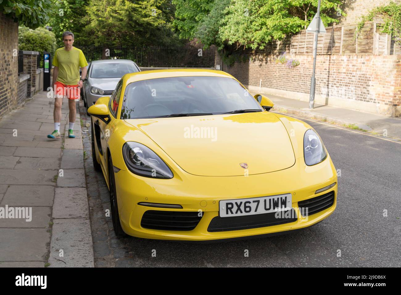 a yellow PORSCHE parks at road side on street parking bay , a jogger with yellow in red short walks toward the car Greenwich London England Stock Photo