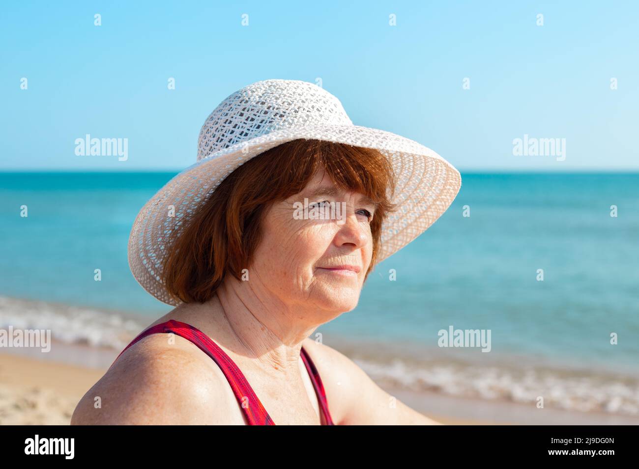 An elderly woman in a hat on the seashore on a sunny day, looks away and smiles, close-up. Summer pastime in retirement. Stock Photo