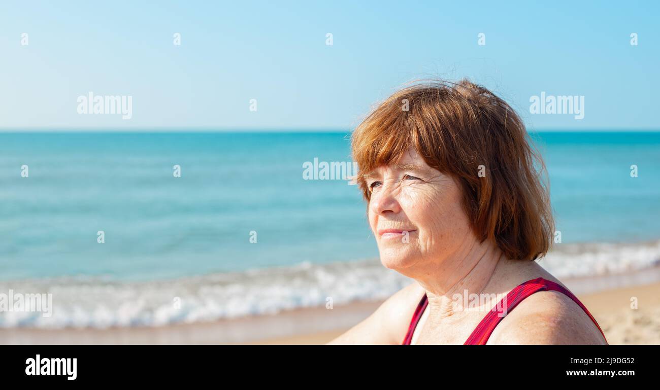 Portrait of a smiling elderly woman sitting on the seashore on a sunny day. Summer pastime in retirement. copy space. Stock Photo