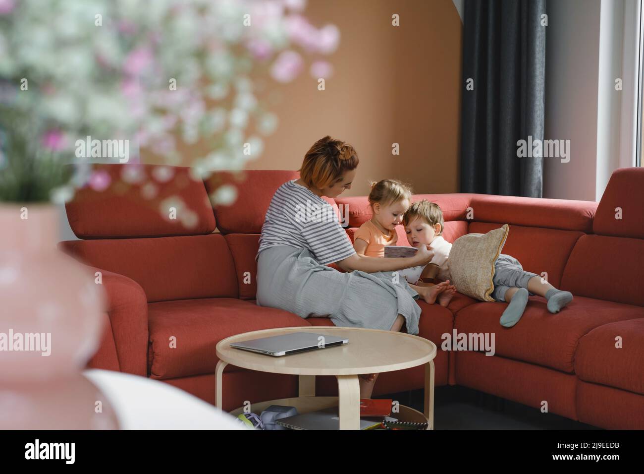 Mother reading book with two kids on sofa. Happy family moments at home. Spending time and educating children. Siblings with mom playing, watching Stock Photo