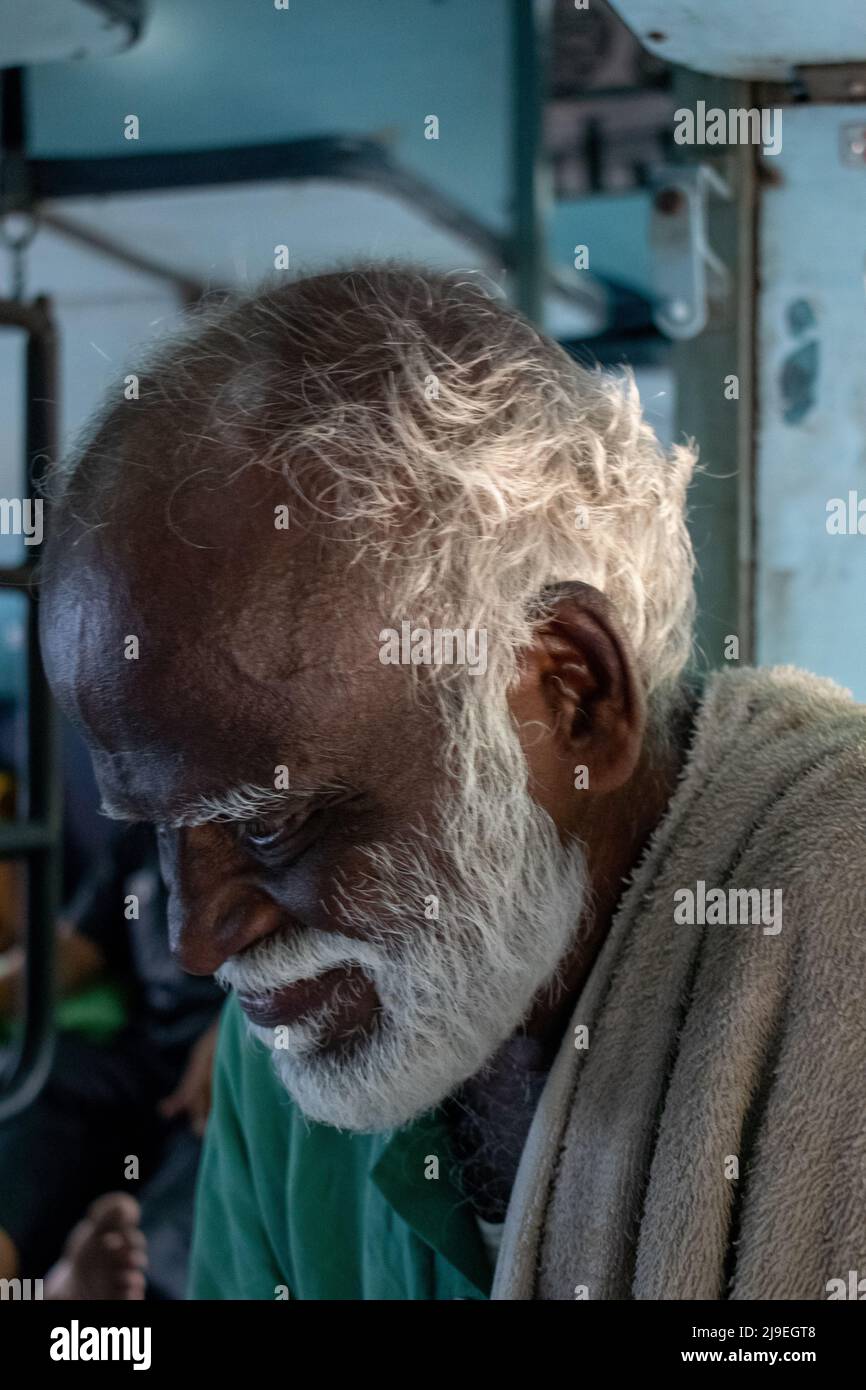 Old senior Indian poor man portrait with a dark brown wrinkled face and white hair and a white beard traveling in train , looks serious. Stock Photo