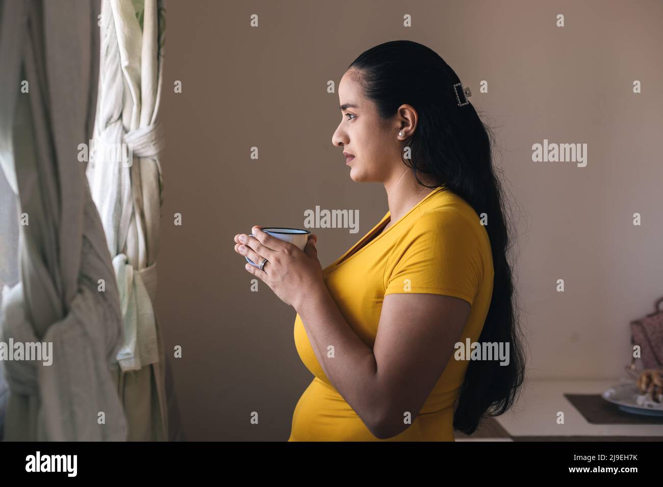 woman in yellow sweater looking out the window with cup in hand Stock Photo