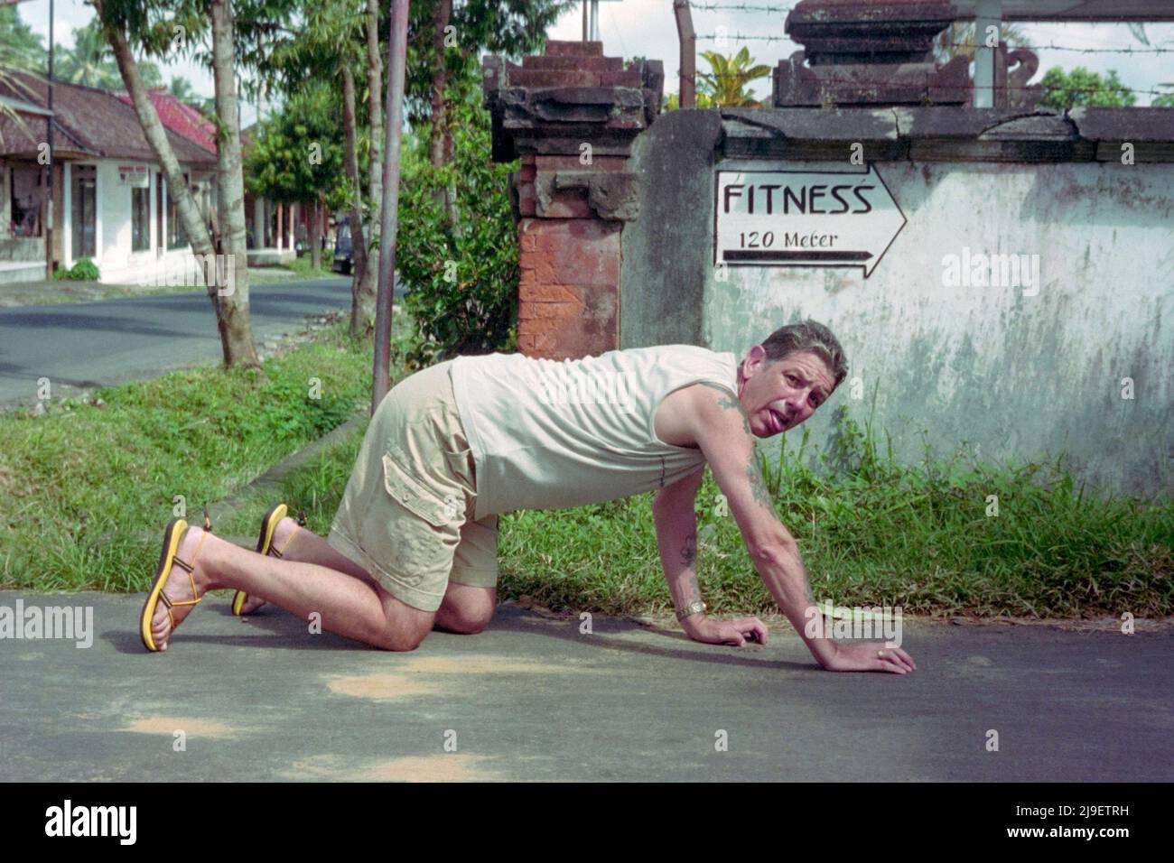 spoof image of exhausted unfit man crawling on hands and knees with sign pointing to fitness centre in background bali indonesia 2004 Stock Photo