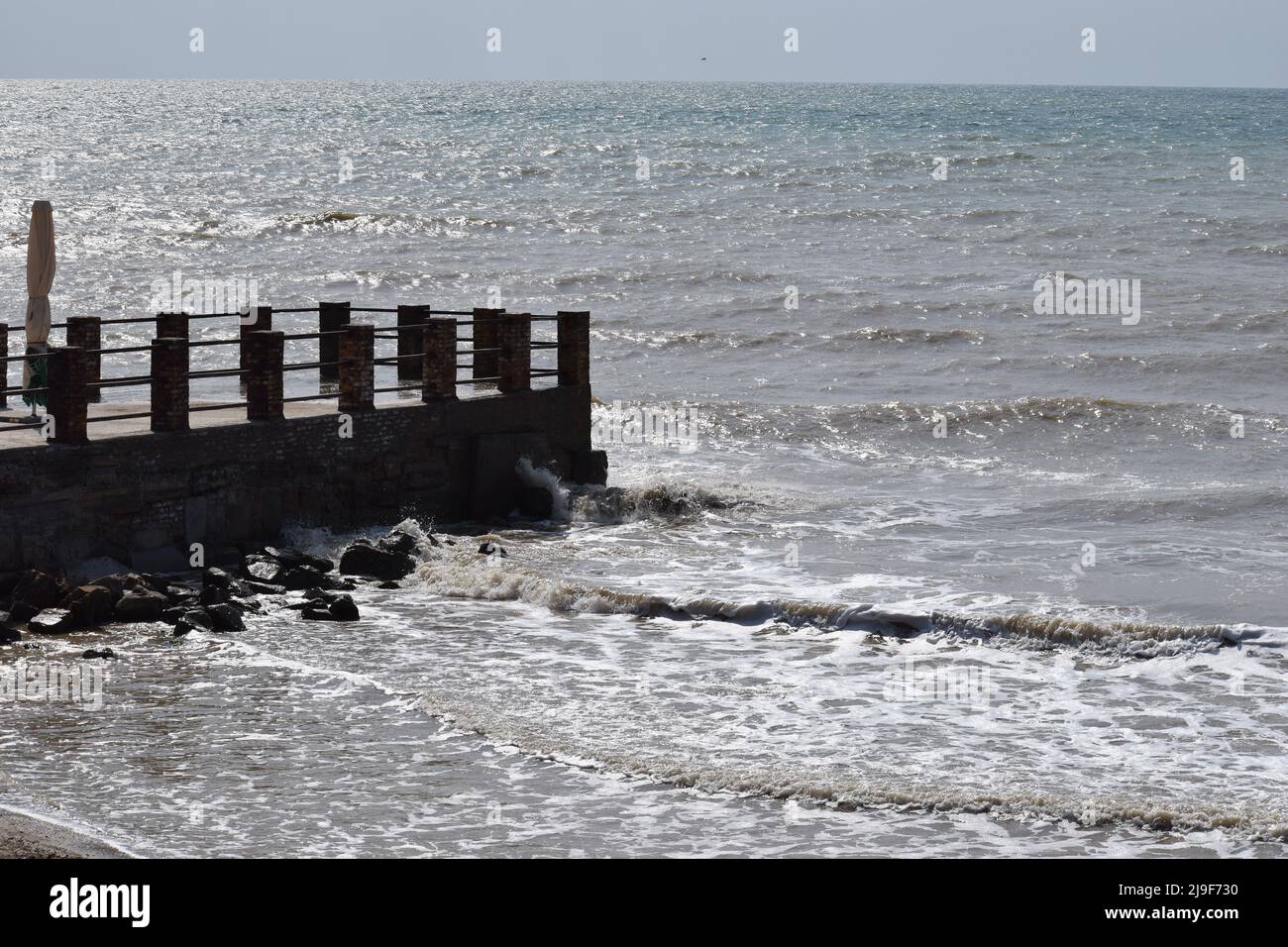 big sea waves splashing against a pier. The pier cuts the sea waves off the coast during a stormy sunny day. Stock Photo