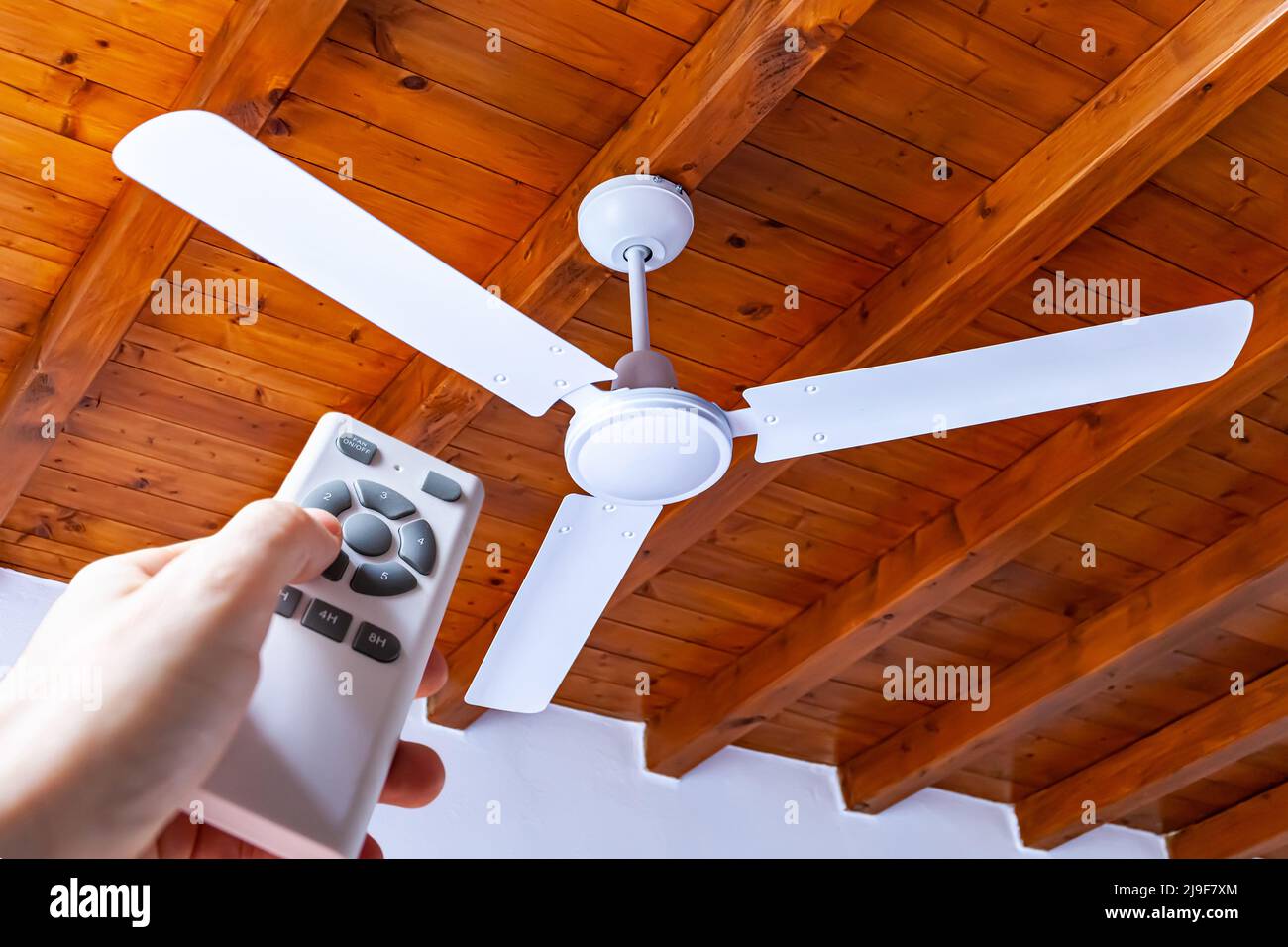 A man uses a remote control to turn on a white ceiling fan mounted in a house with wooden ceilings. Stock Photo
