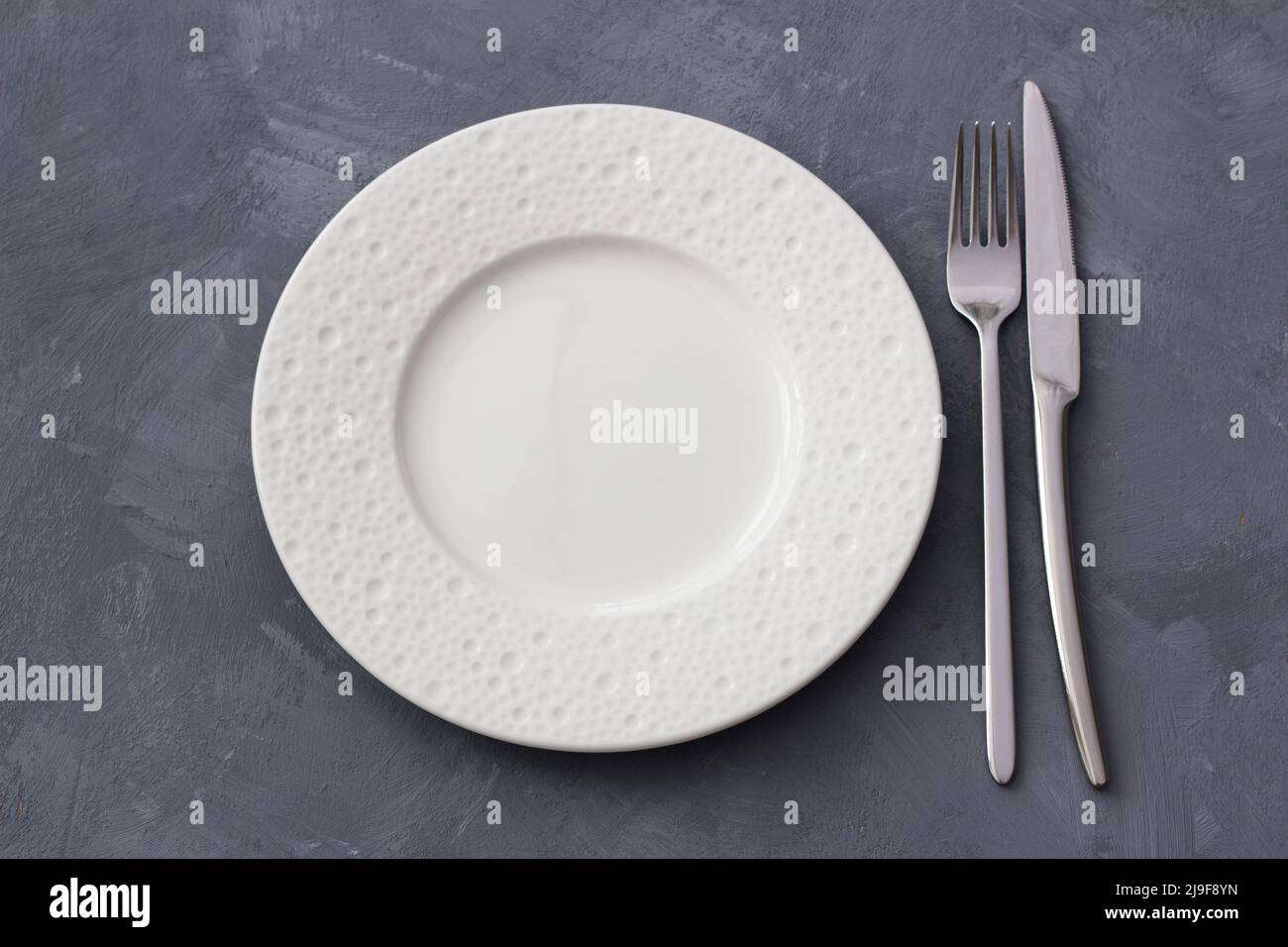 White ceramic plate on a gray textured background with fork and knife ...