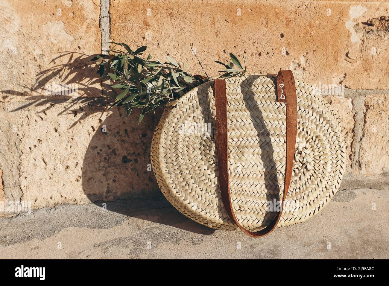 Summer vacation still life. Round French straw basket with leather handles and green olive tree leaves and branches in sunlight. Golden sandstone wall Stock Photo