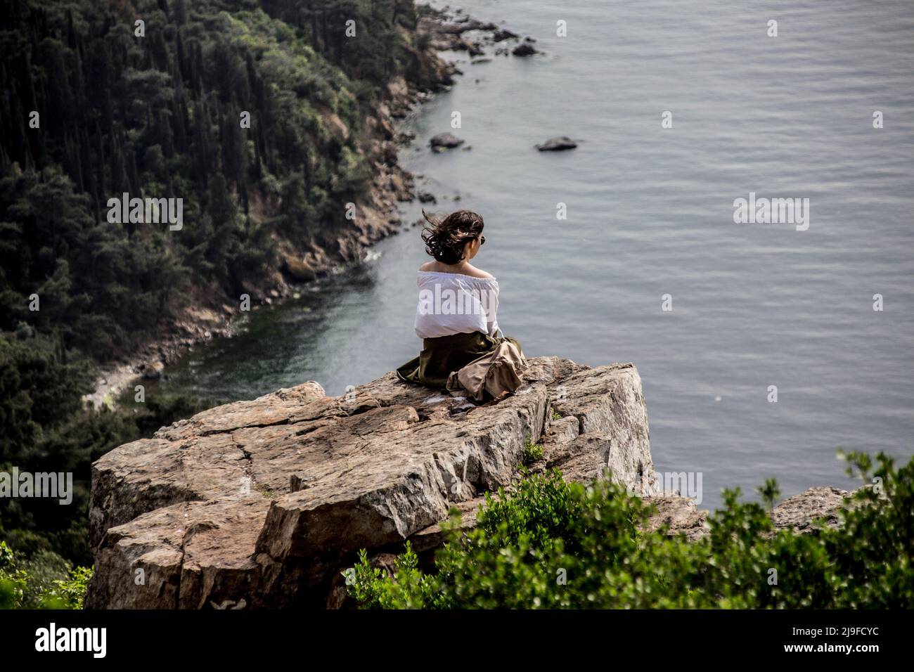 Young girl sitting on the edge of the cliff, on the edge of the cliff, in Büyükada, Istanbul, at the sea bay in the forest, a person sitting on a ston Stock Photo