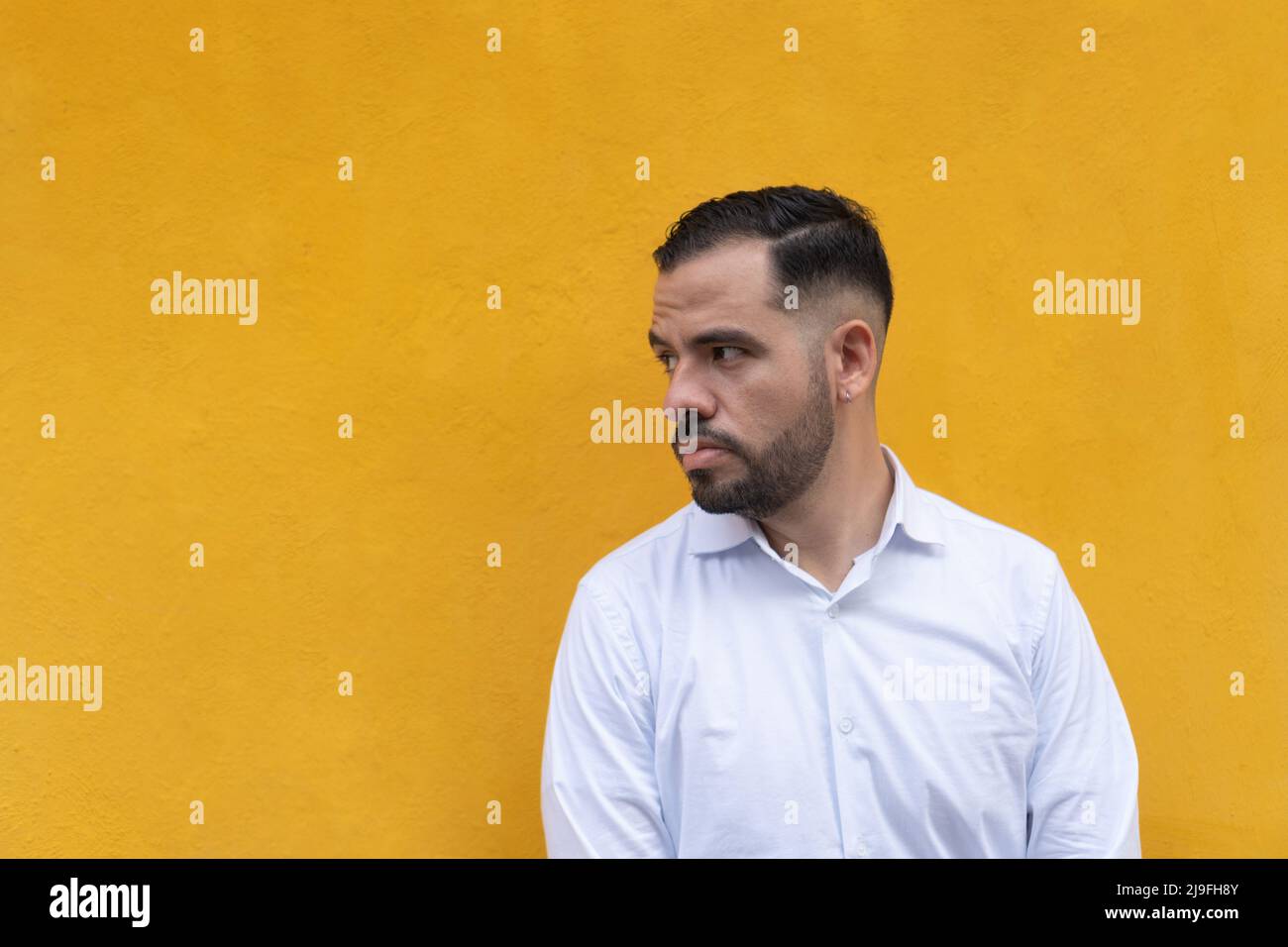 Latin man standing in front of a yellow wall. He is looking to the side with seriousness Stock Photo