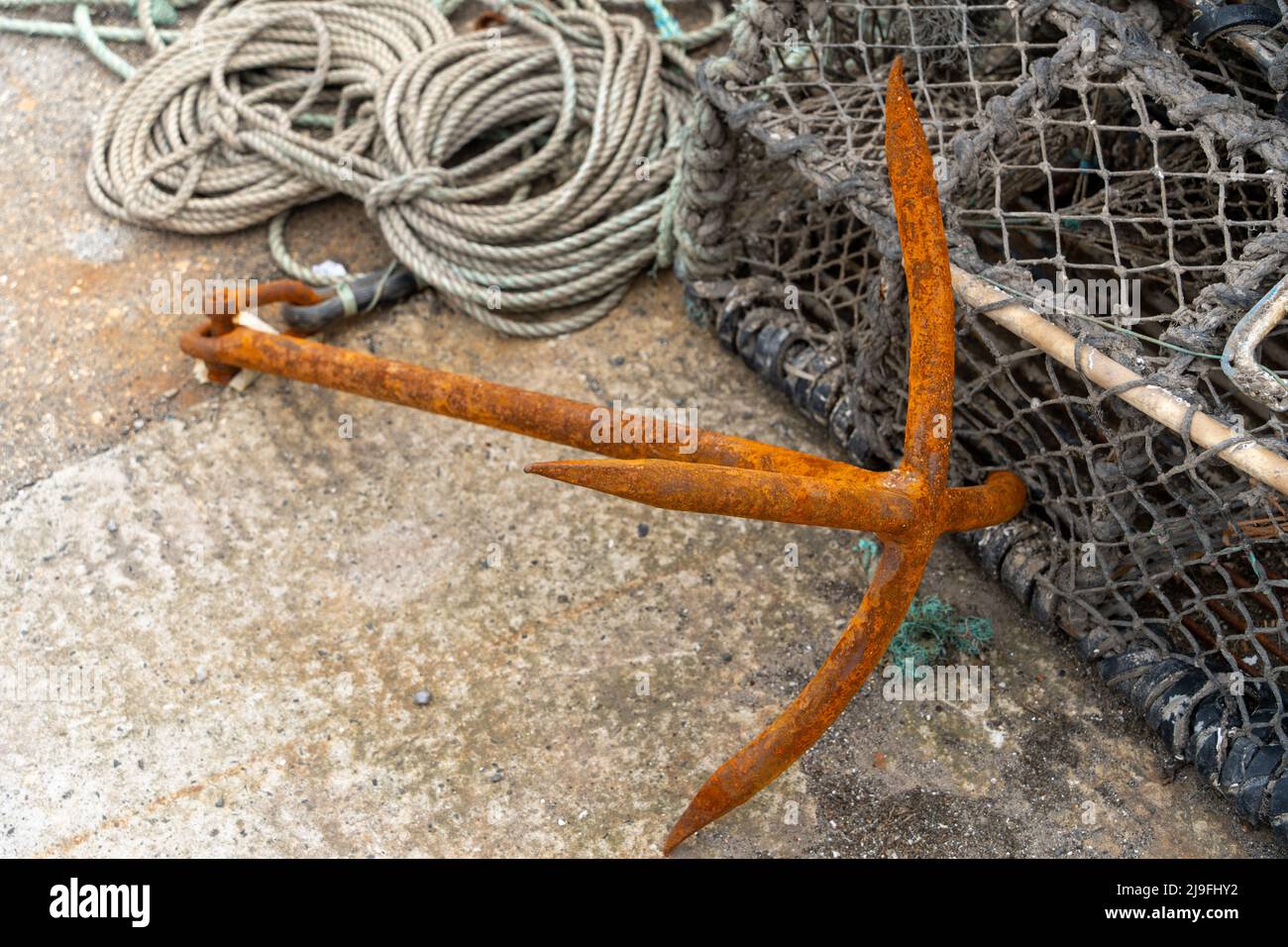 A rusty anchor next to a crab or lobster pot and a coil of rope, used in the fishing industry.  Concept of being anchored. Stock Photo